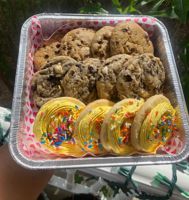 A foil tray of chocolate chip and cookies and cream cookies with frosted sprinkle cookies, lined with heart paper